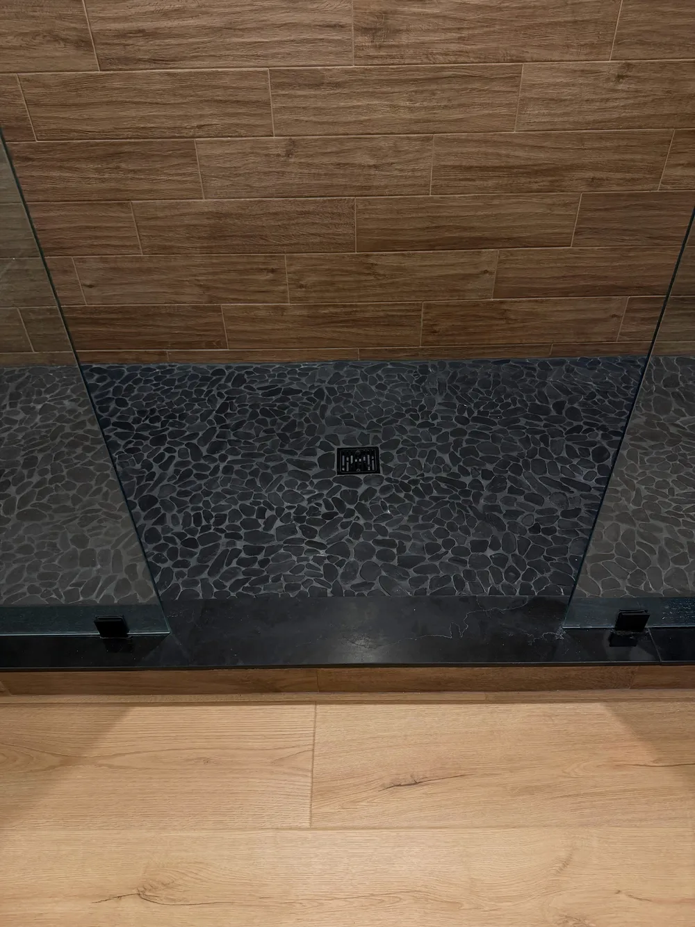 Dark pebble-style shower floor, center drain, black threshold, and adjoining light wood-look flooring.
