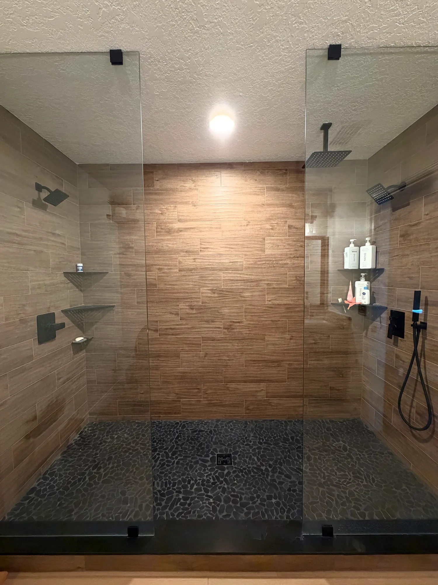 Expanded glass-enclosed shower with two shower stations, warm wood-look wall tile, and a dark pebble-style floor.