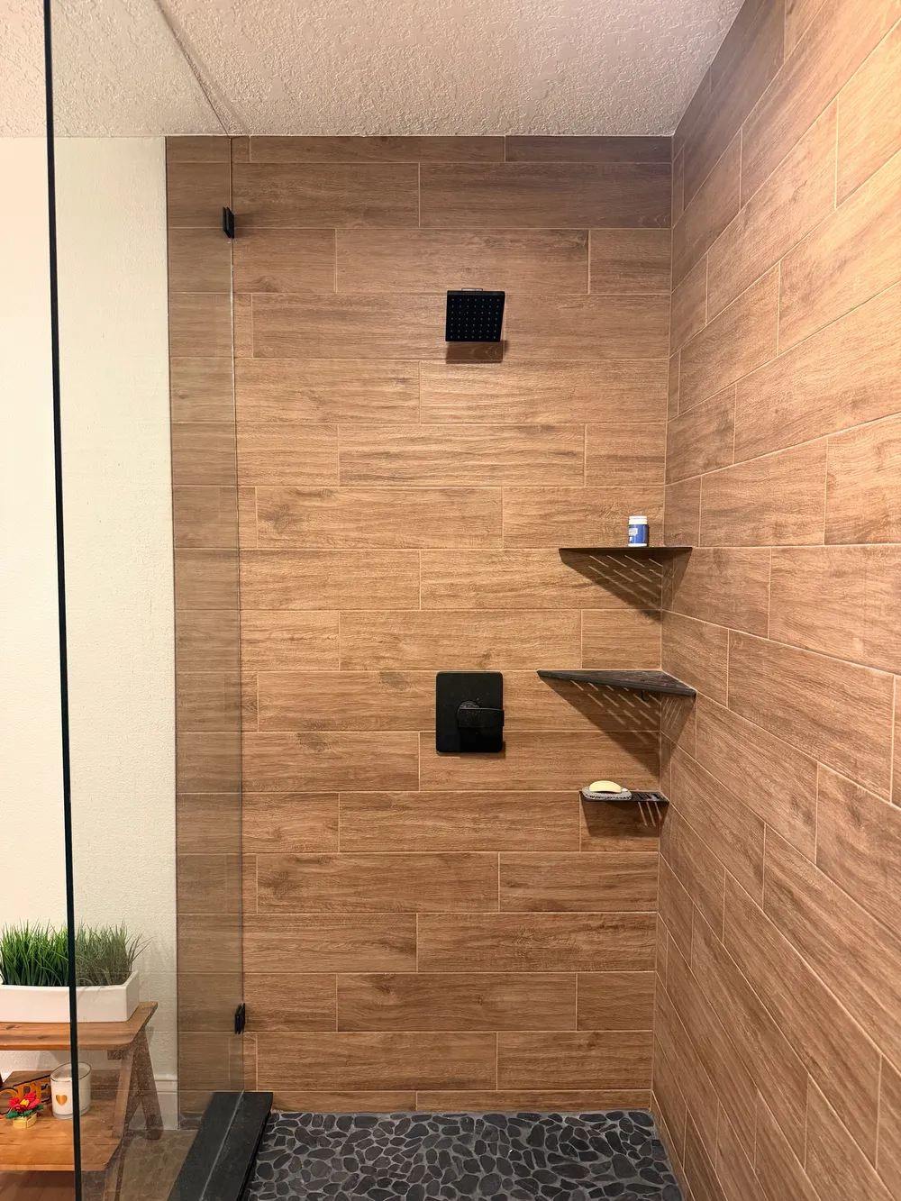 Wood-look shower wall tile with a black showerhead, control, and coordinating metal corner shelves.