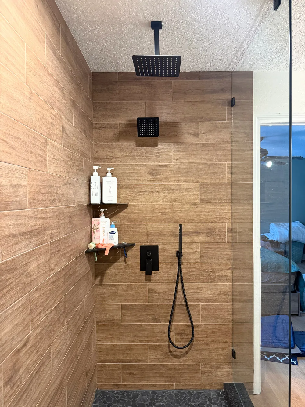 Second shower station with black rainfall and handheld fixtures, corner shelving, and wood-look wall tile.