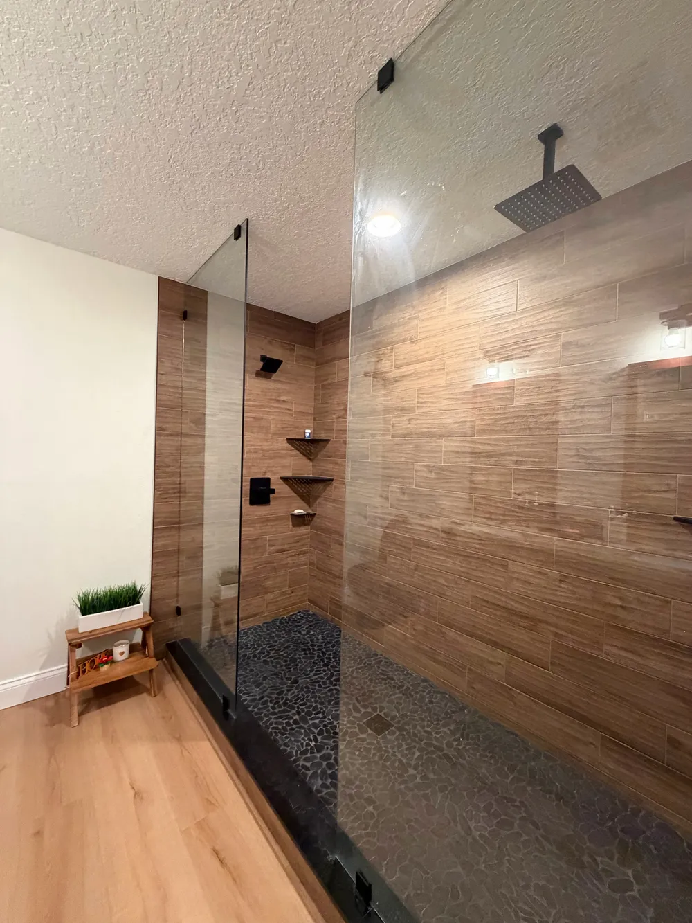 Angled view of the frameless shower enclosure, wood-look wall tile, black fixtures, and light bathroom flooring.