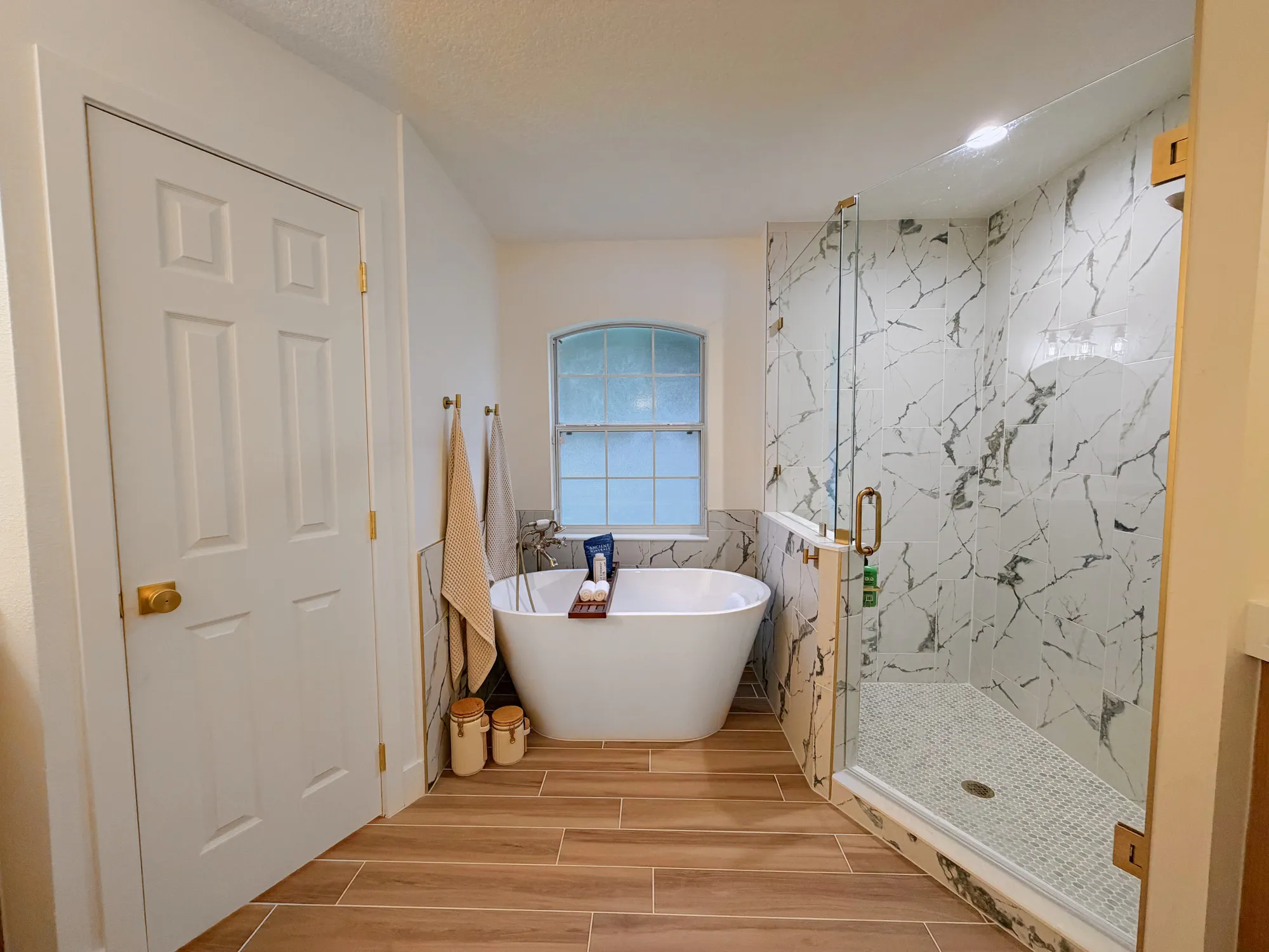 Finished Calle bathroom with a freestanding tub, glass-enclosed shower, marble-look tile and warm wood-look flooring.