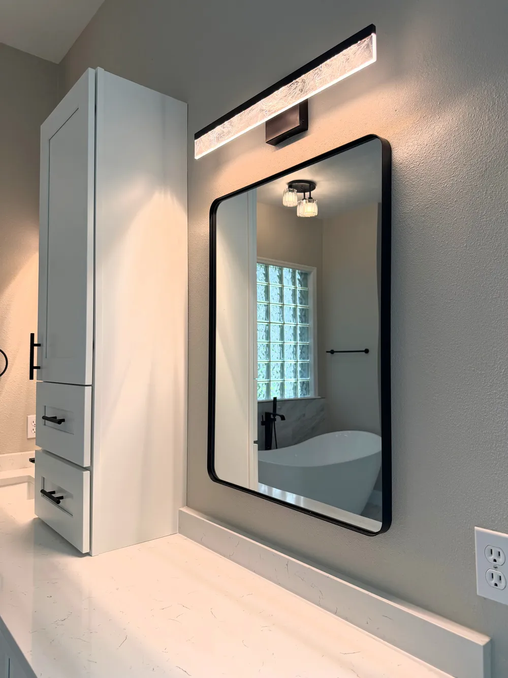 White storage tower and black-framed mirror above the Estancia vanity, lit by a warm wall fixture.