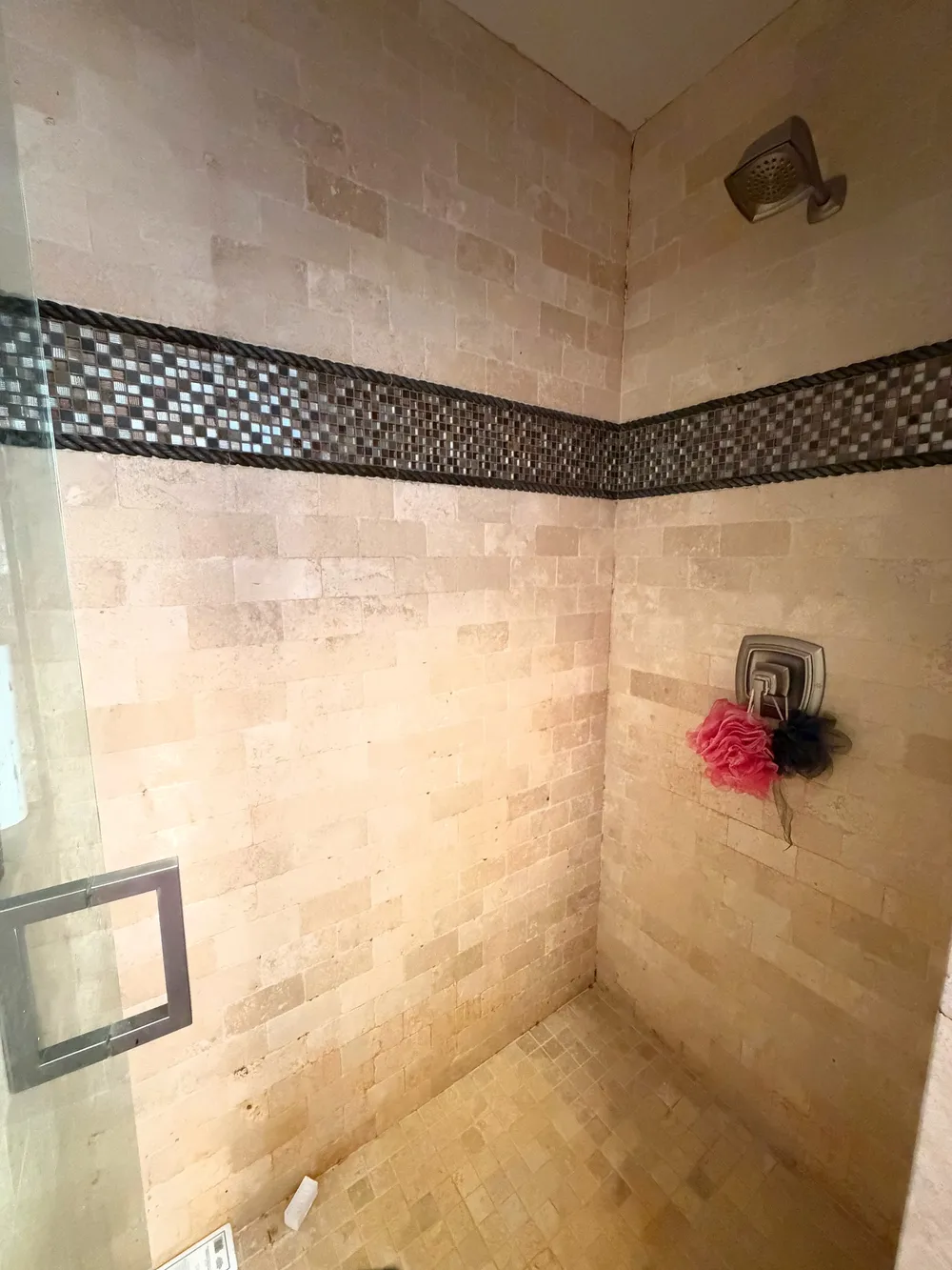 Original enclosed shower with warm stone-look tile and a dark mosaic band.