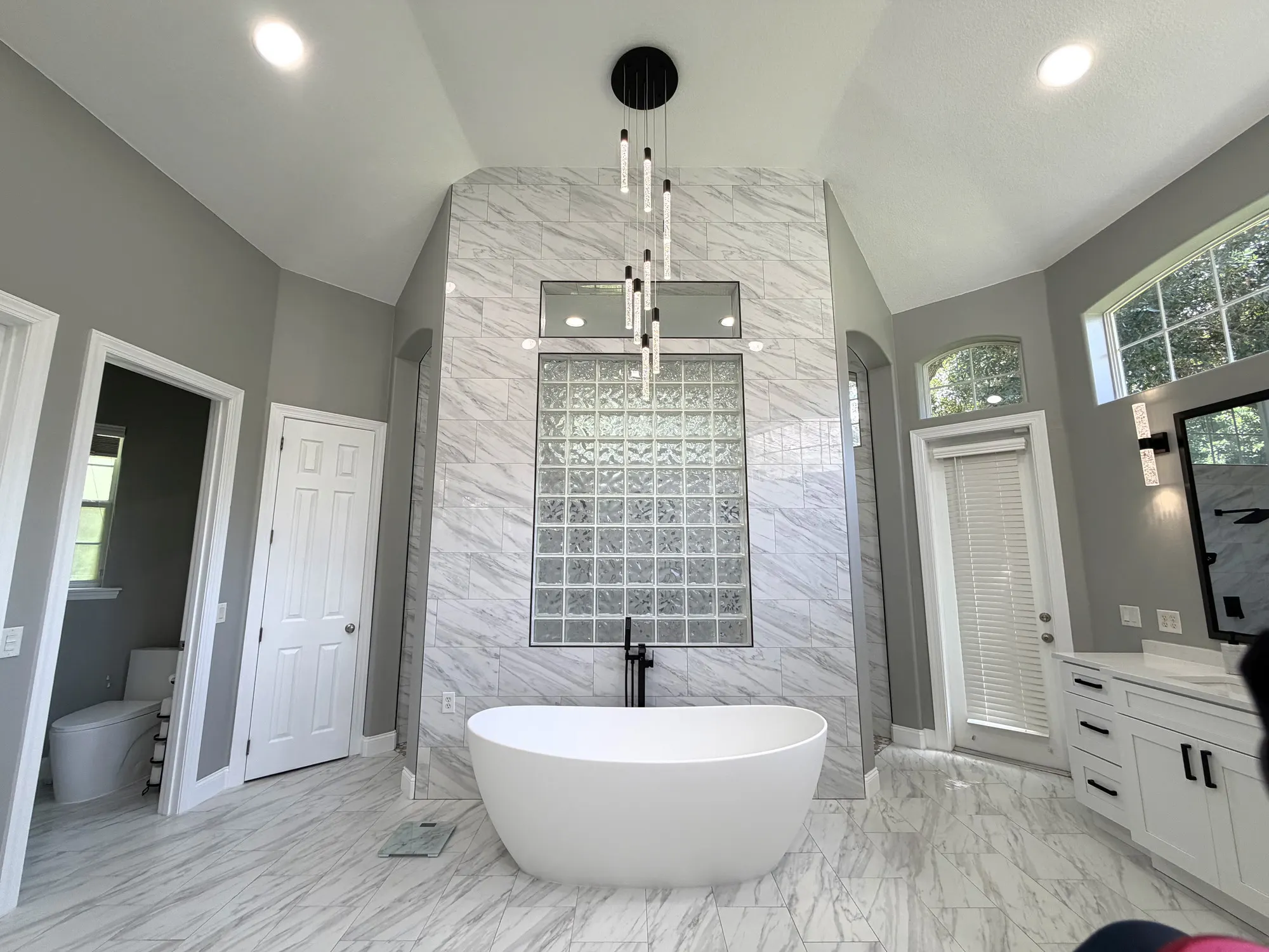 Freestanding soaking tub beneath a glass-block wall and full-height veined tile in The Grand Revival bathroom.
