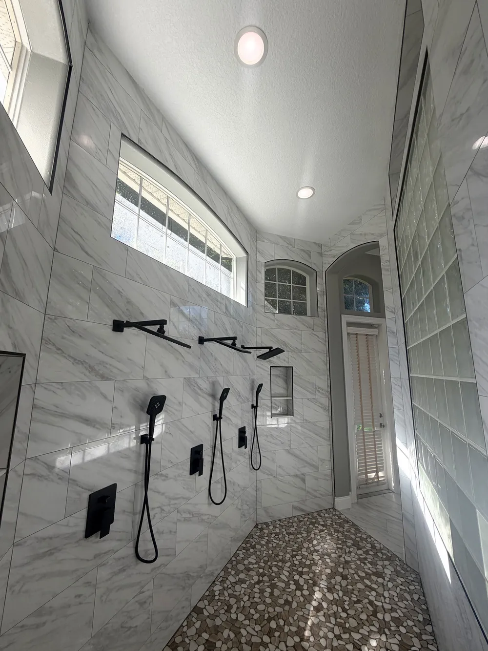 Tall tiled shower with two black rainfall showerheads, handheld fixtures and a pebble-style floor in The Grand Revival bathroom.
