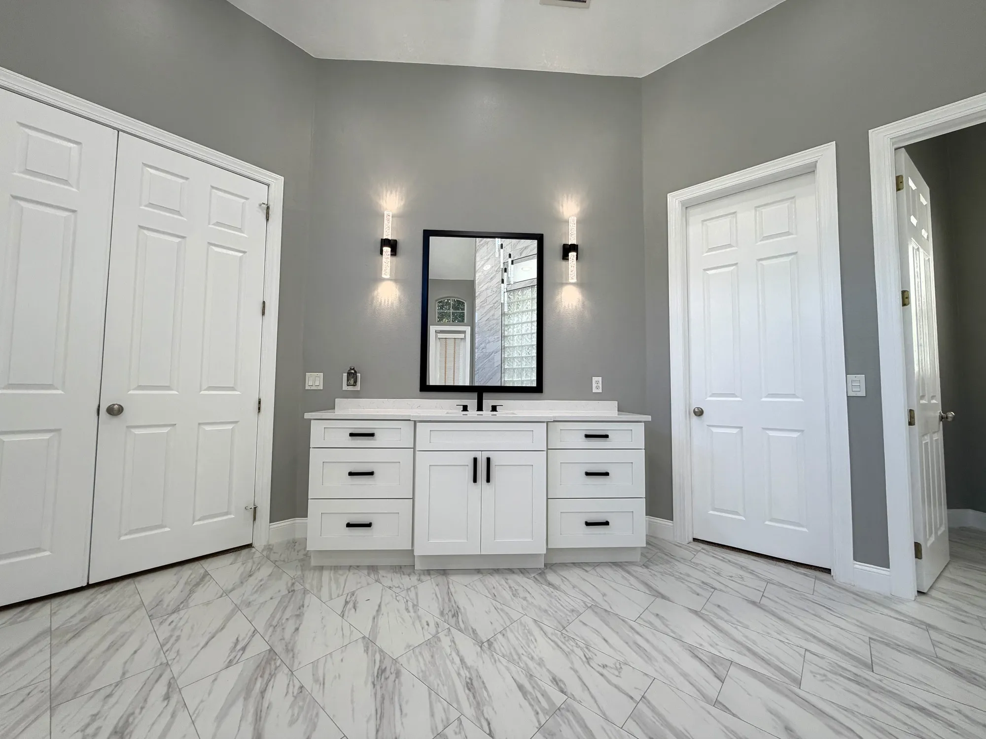 White shaker vanity with a black-framed mirror and paired wall sconces between two doors in The Grand Revival bathroom.