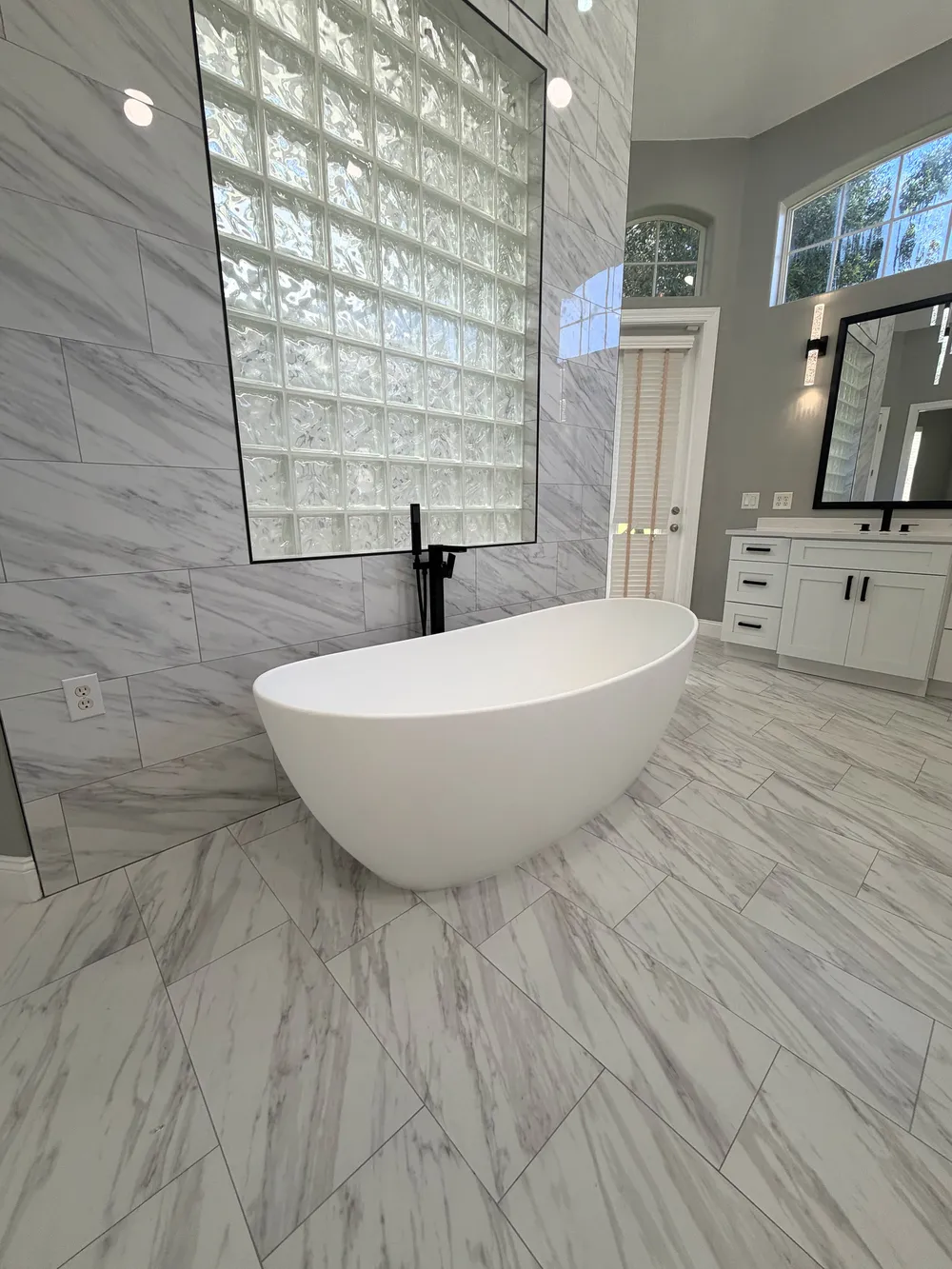 Freestanding white tub and matte-black floor-mounted faucet below the preserved glass-block wall in The Grand Revival bathroom.