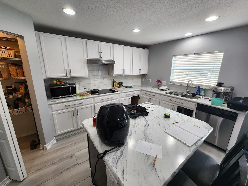 Earlier kitchen with white cabinets, a metal range hood, subway tile, double-basin sink, and narrow island; lower cabinet doors have been removed.