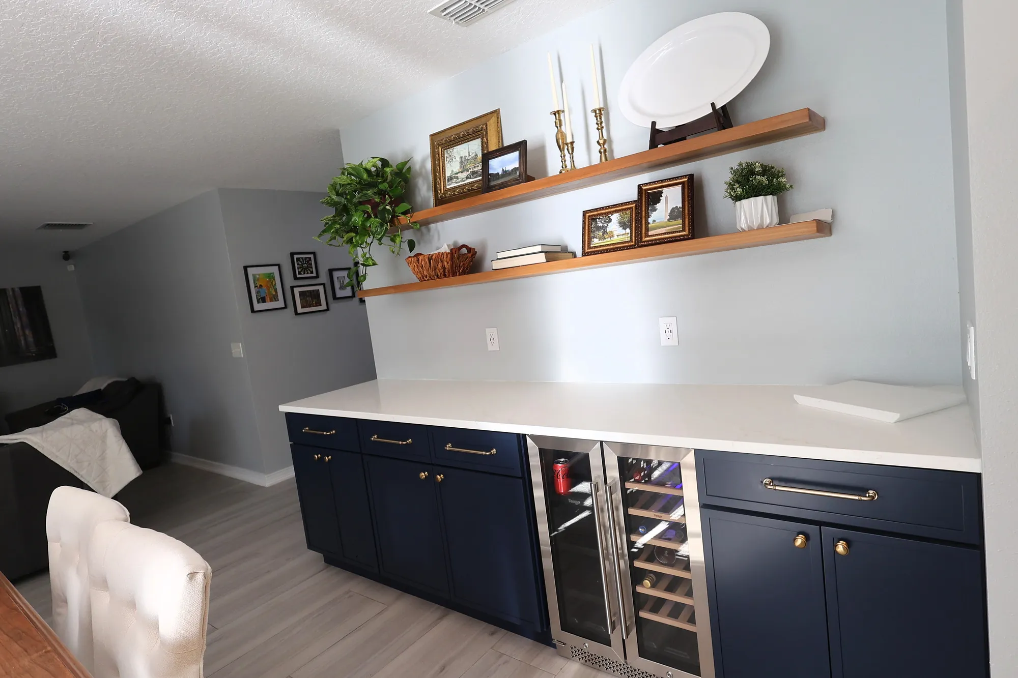 Separate dry bar with dark blue cabinetry, brass hardware, a glass-front beverage cooler, quartz counter, and wood shelves.