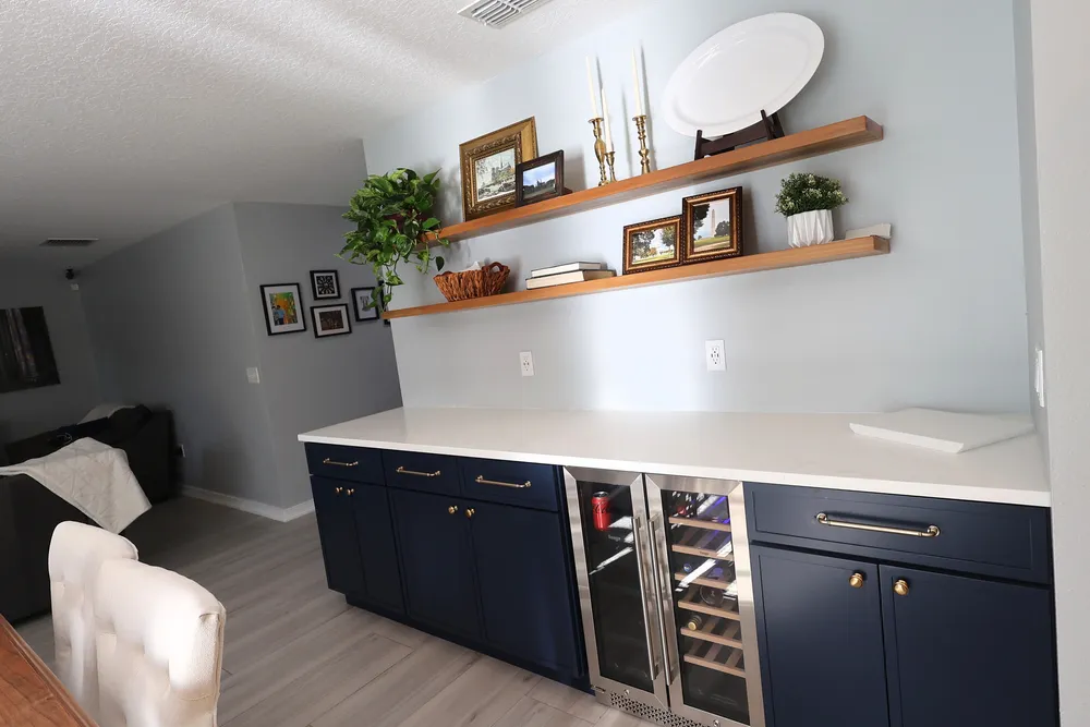 Separate dry bar with dark blue cabinetry, brass hardware, a glass-front beverage cooler, quartz counter, and wood shelves.