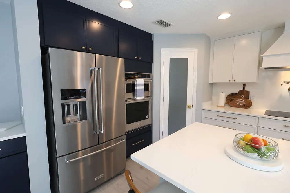 Stainless refrigerator and wall oven surrounded by dark blue cabinetry with brass knobs, beside the white quartz island.