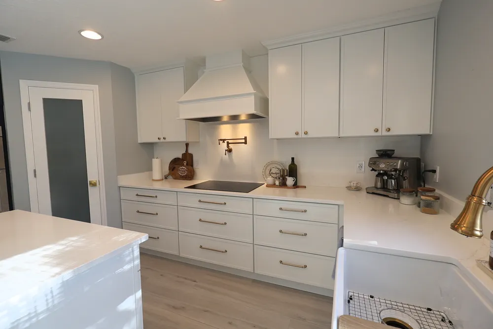 Finished cooking wall with a decorative range hood and brass pot filler, beside a white farmhouse sink and quartz counters.