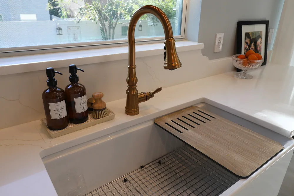 White farmhouse sink with metal basin grids, a brass bridge faucet, and veined quartz counters beneath the kitchen window.