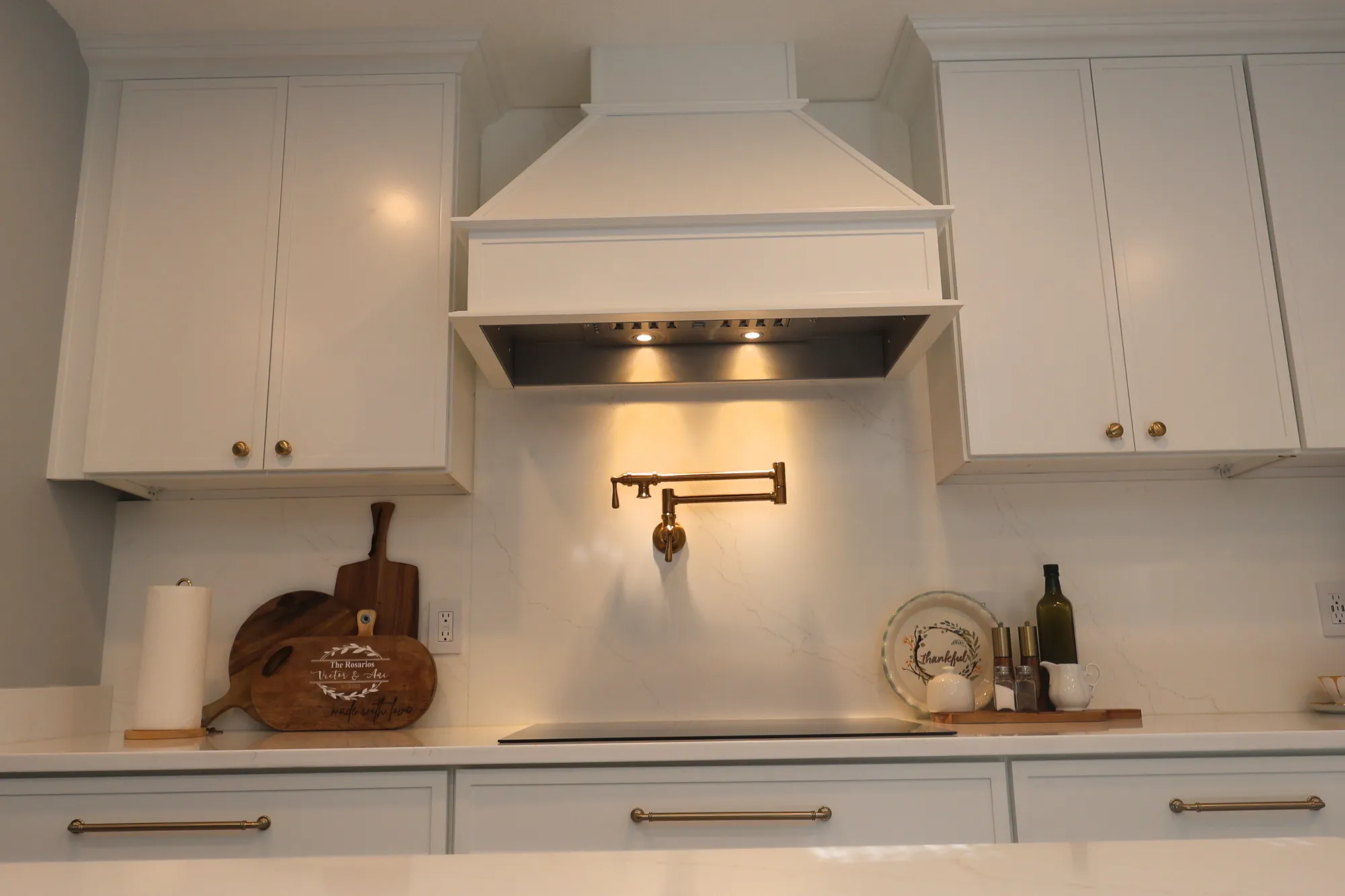 Decorative white range hood above a brass pot filler, quartz backsplash, and cooktop, with warm task lighting.