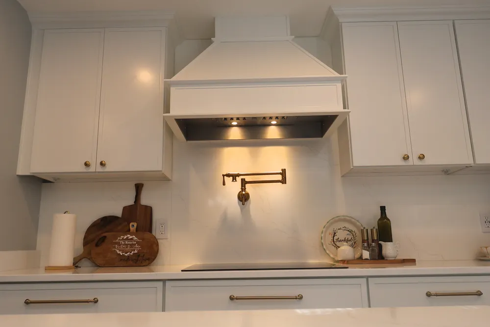 Decorative white range hood above a brass pot filler, quartz backsplash, and cooktop, with warm task lighting.